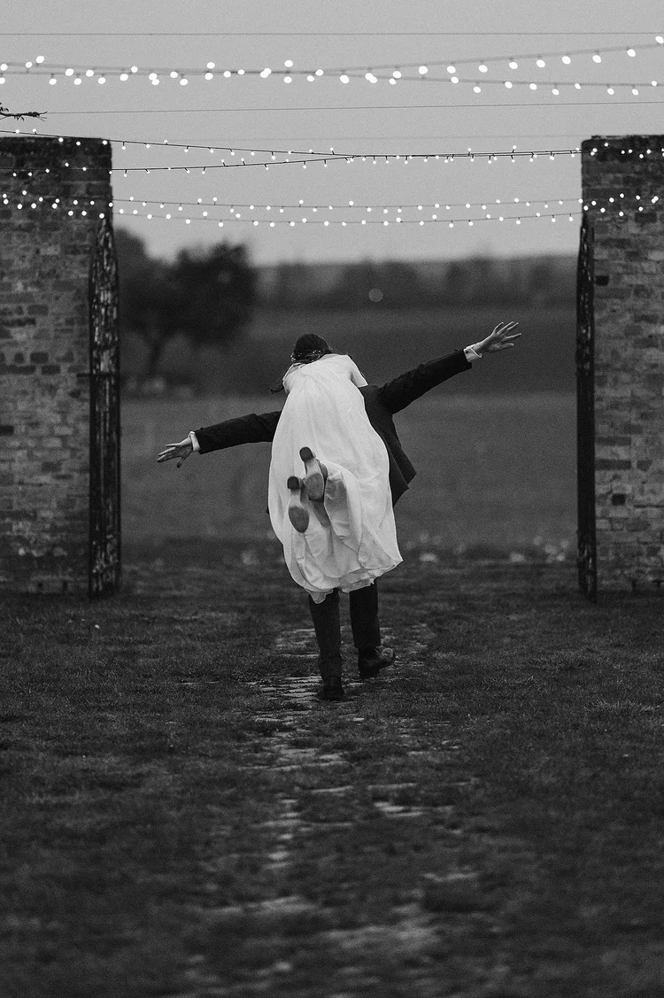 Groom carrying the bride beneath string lights on a rainy wedding day